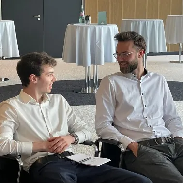 Zwei junge Männer in weißen Hemden sitzen nebeneinander und unterhalten sich, im Hintergrund Stehtische mit weißen Hussen.