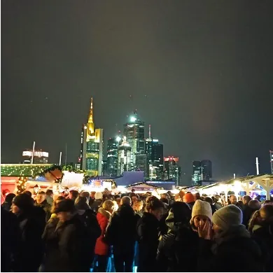Abendlicher Weihnachtsmarkt mit vielen Besuchern, im Hintergrund die beleuchtete Hochhaus-Skyline von Frankfurt am Main.