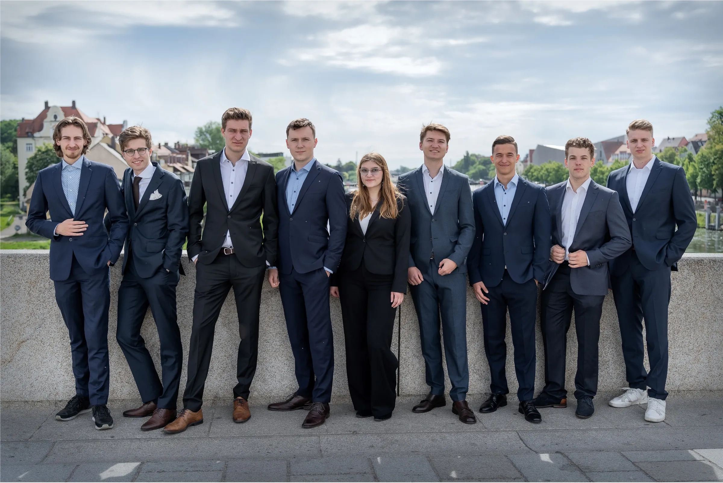 Gruppenfoto des Vorstands 2023–2024: neun Personen in dunklen Anzügen lehnen nebeneinander an einer steinernen Brüstung, dahinter ein Fluss und Häuser am Ufer.